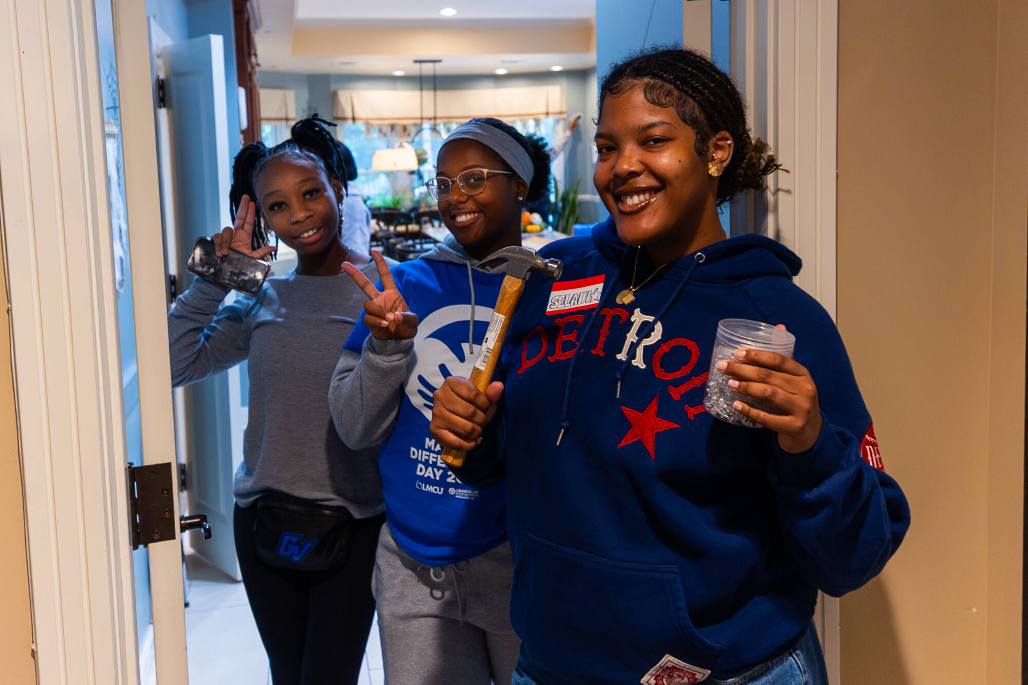 Three young women stand in a doorway; one holds a hammer, another a container of nails, while the third makes a peace sign, all wearing casual gear.